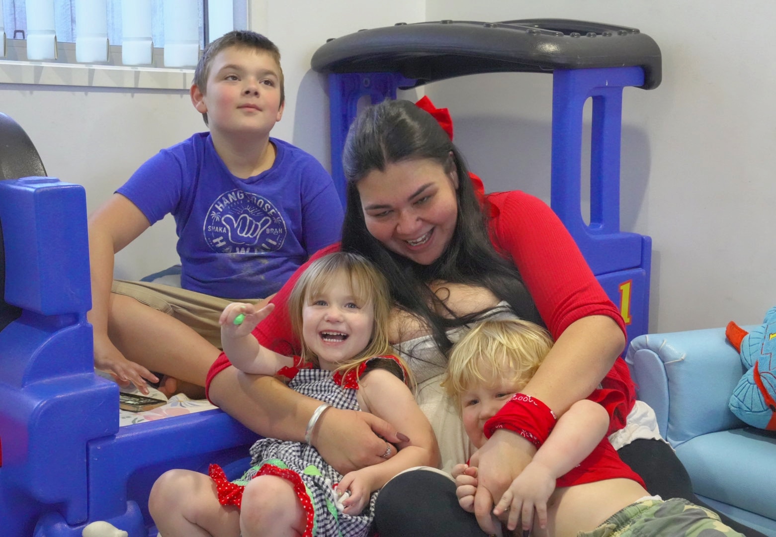 A woman and her three children sit on the floor in a room laughing and hugging.