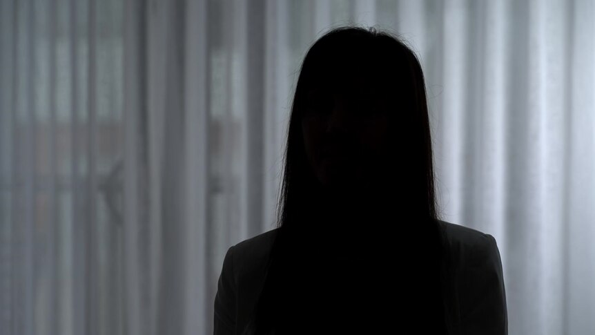 The darkened silhouette of a woman with straight long hair against a non-descript white curtain.