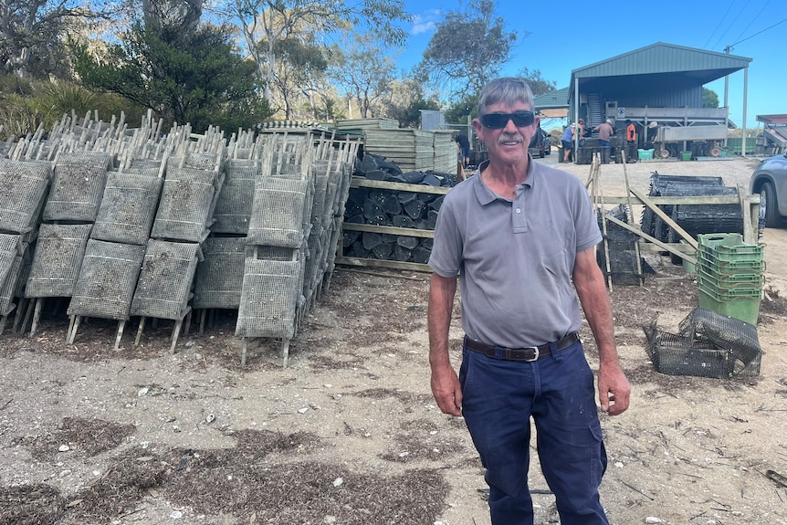 a man stands in front of oyster racks piled up on a sandy beach