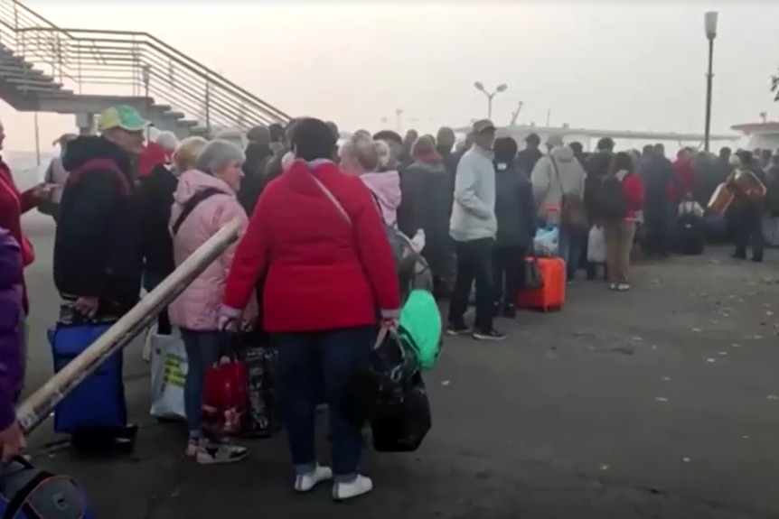 A long queue of people in winter clothing and holding bags at an industrial-looking dock.