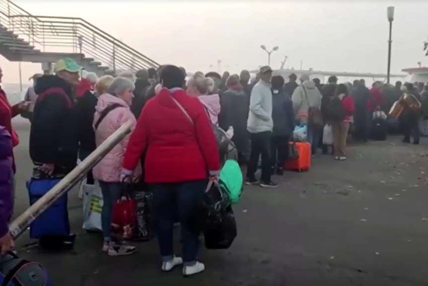 Una lunga fila di persone vestite con abiti invernali che trasportano borse su un marciapiede dall'aspetto industriale.