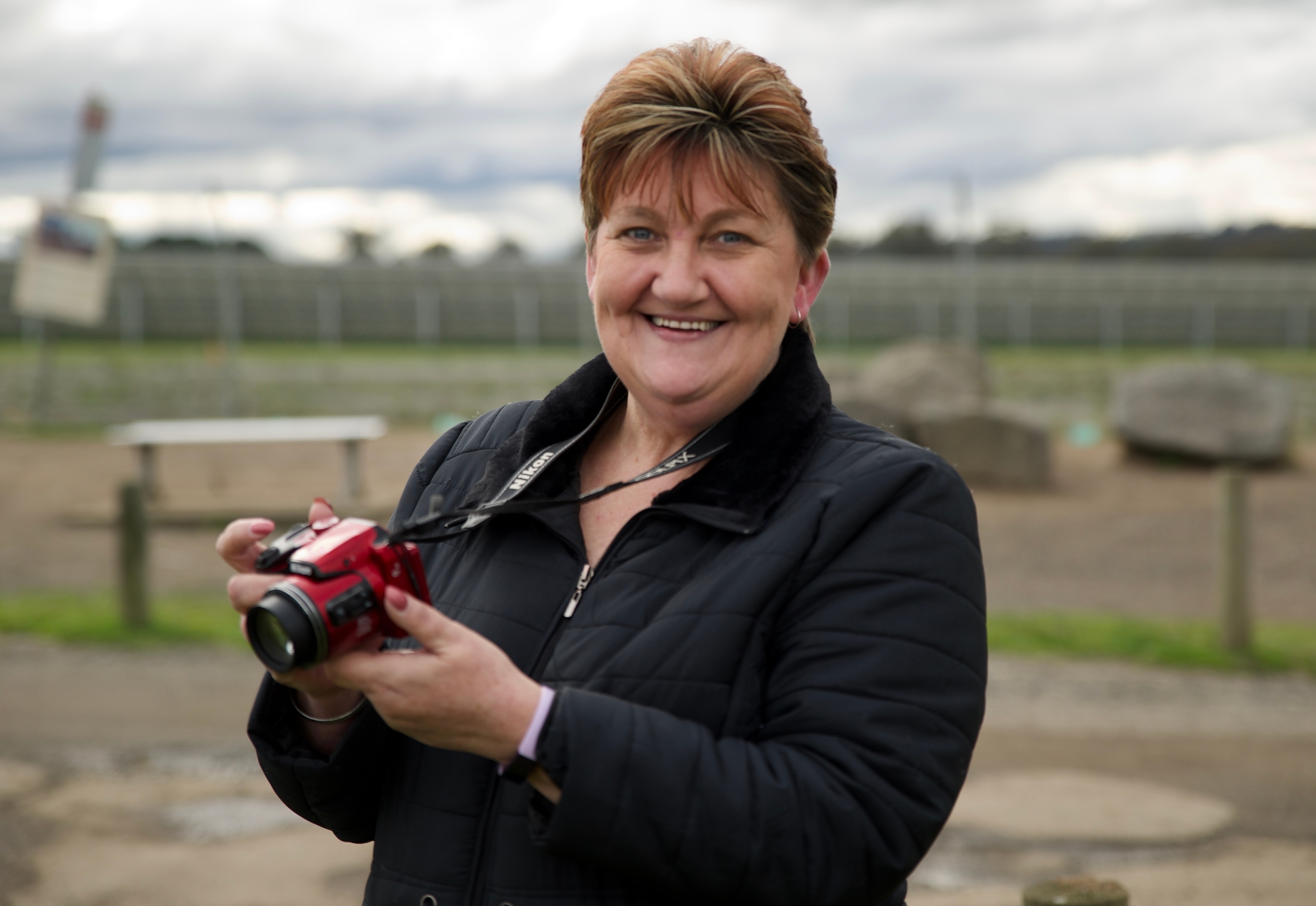 a woman with short hair holding a red camera.  She is smiling and wears a black jacket.