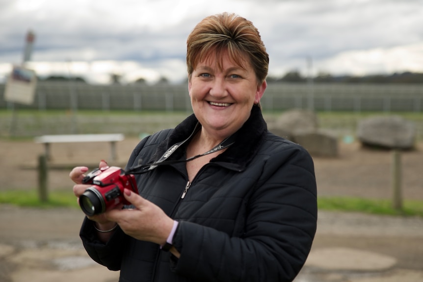 a woman with short hair holding a red camera.  She is smiling and wears a black jacket.