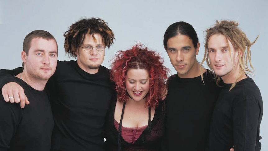 A red-haired woman, three men, two with blond and dark-haired  dreadlocks, all in black stand close, smile.