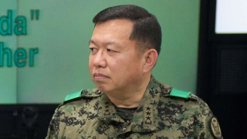Man in military fatigues stands in a room with a green wall, looking to his right with a neutral expression