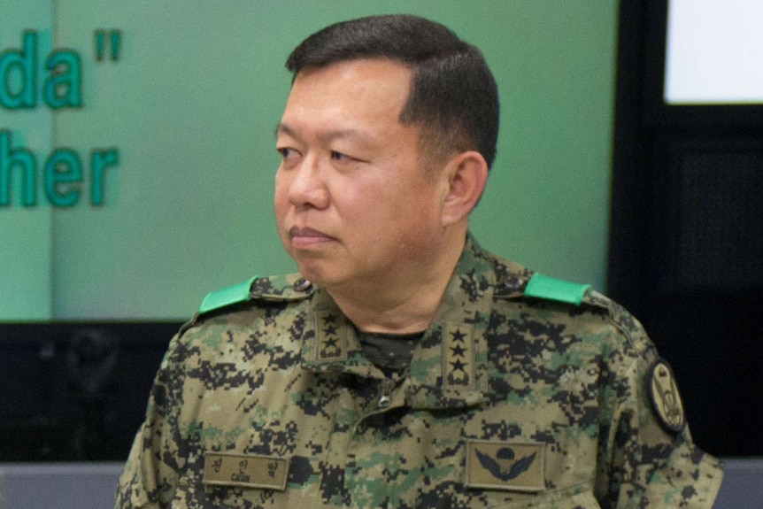 Man in military fatigues stands in a room with a green wall, looking to his right with a neutral expression