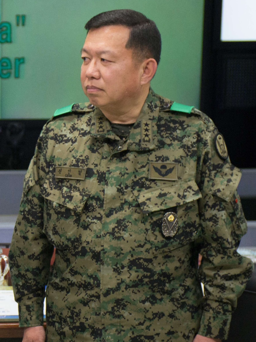 Man in military fatigues stands in a room with a green wall, looking to his right with a neutral expression