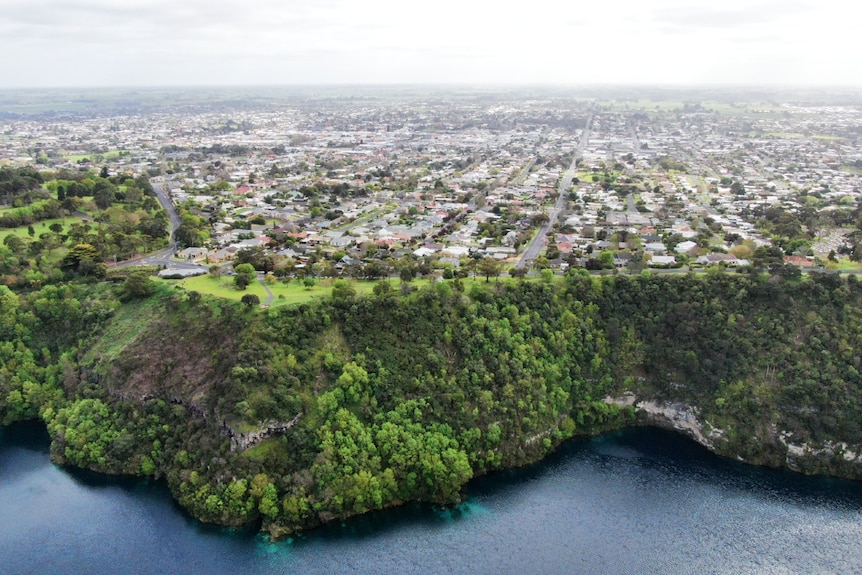 A blue lake, with a green cliff and a rural city stretching on behind the cliff.