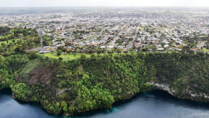 A blue lake, with a green cliff and a rural city stretching on behind the cliff.