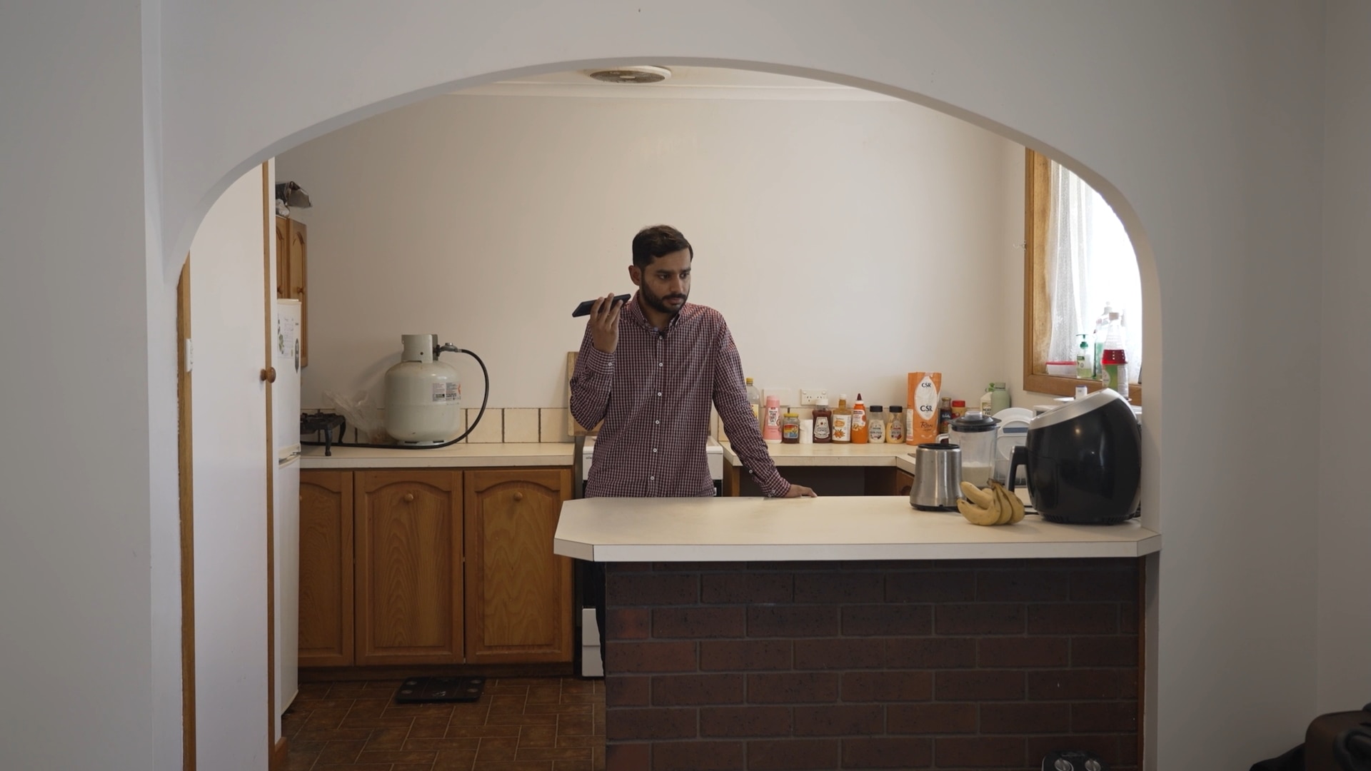 A man with combed black hair and stubble in a shirt stands in his kitchen holding his phone to listen to a voice note.