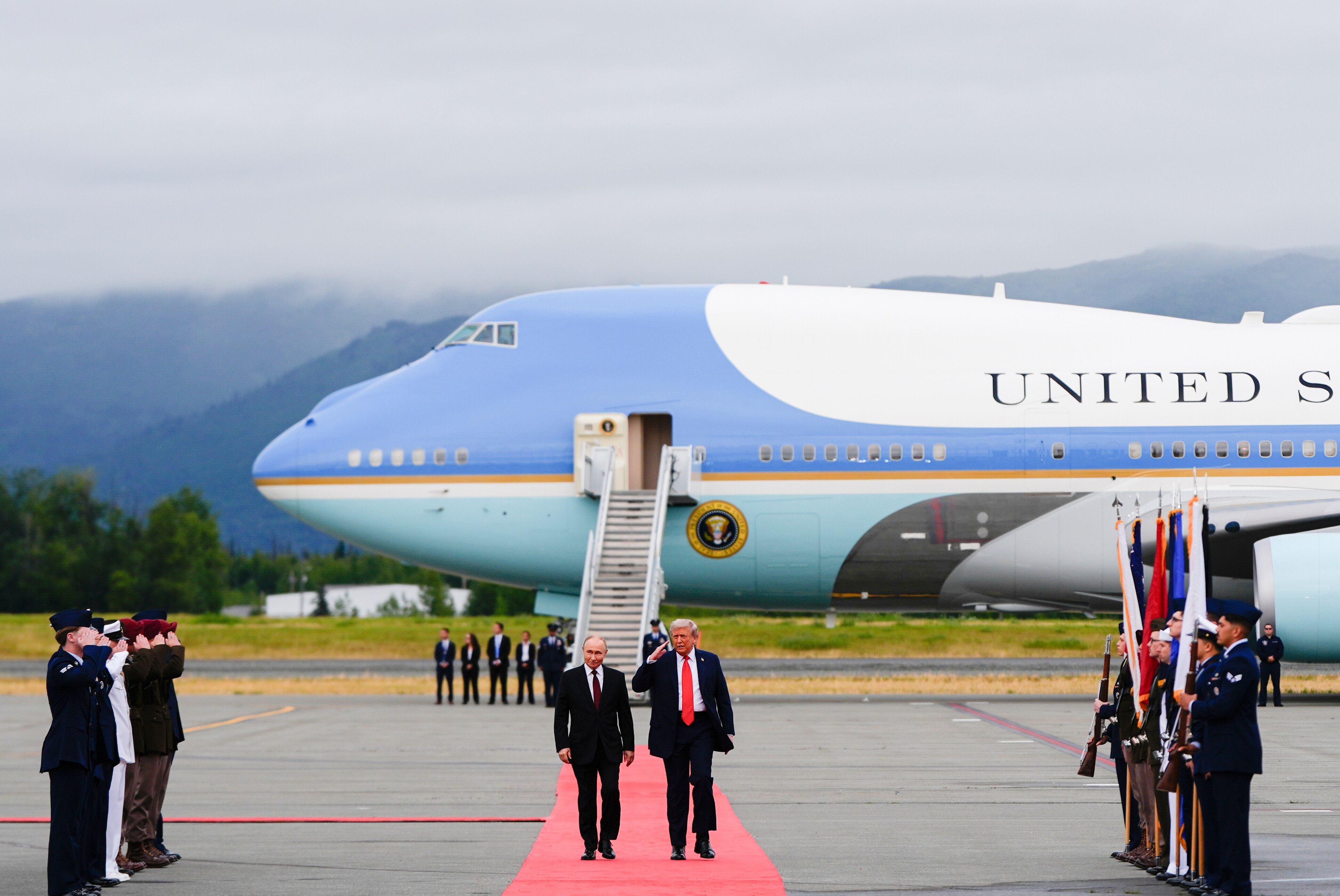 A long shot of Trump and Putin walking a red carpet, being saluted by officials holding flags.