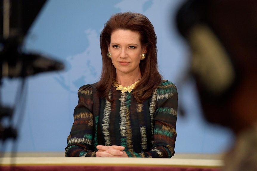 Still from a tv show, a female newsreader in 80s style sits as a desk on a tv set