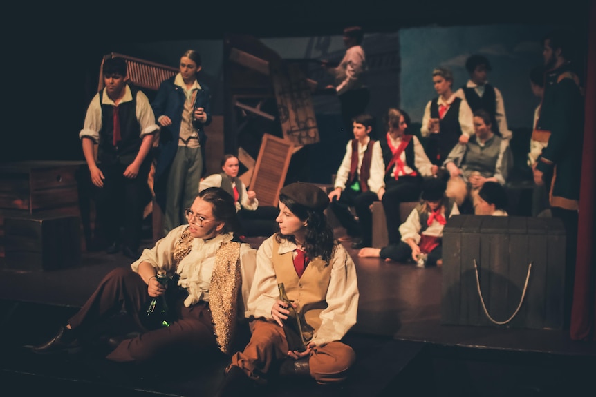 A group of young people dressed in waist coats and white shirts, on stage surrounded by wood boxes.