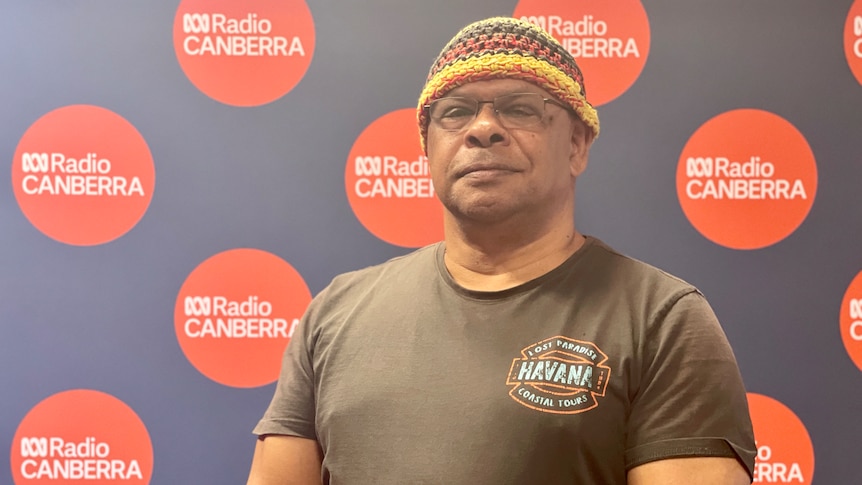 An indigenous man wearing a yellow, red and black beanie and black tshirt, standing proudly, looking at the camera.