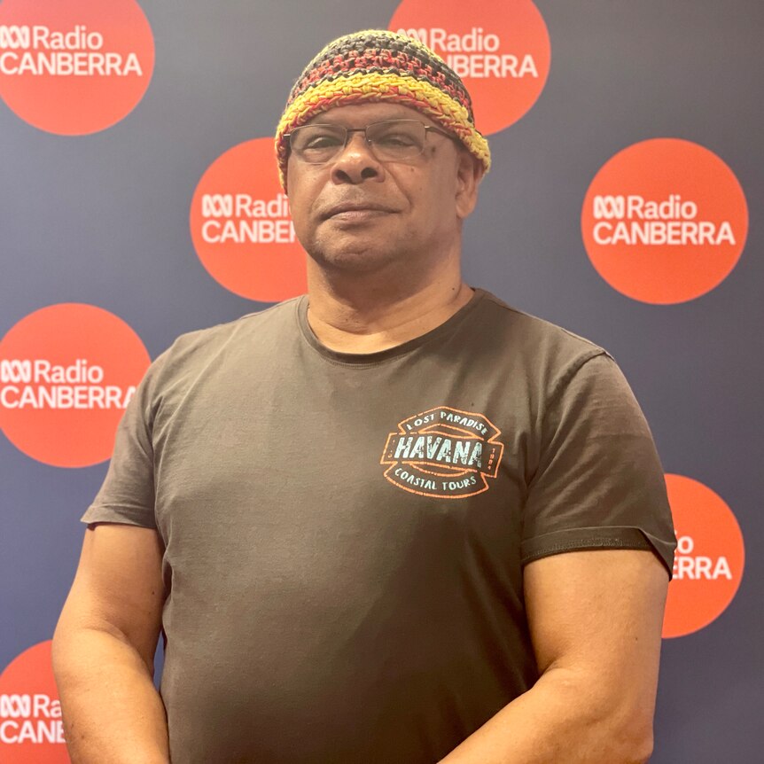 An indigenous man wearing a yellow, red and black beanie and black tshirt, standing proudly, looking at the camera.