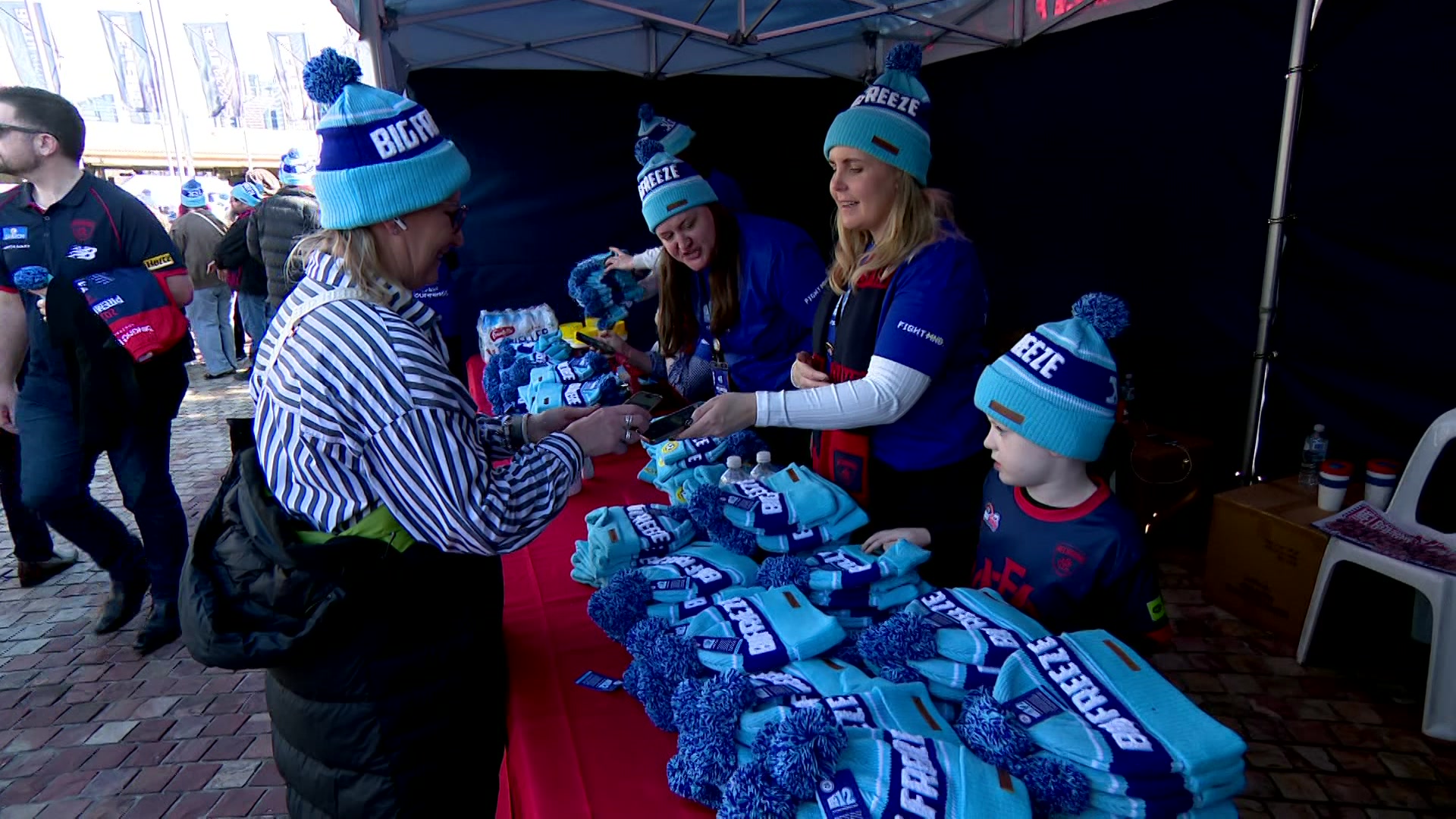 Two women and a child in blue beanies stand behind a counter stacked with beanies and serve a woman wearing the same beanie.