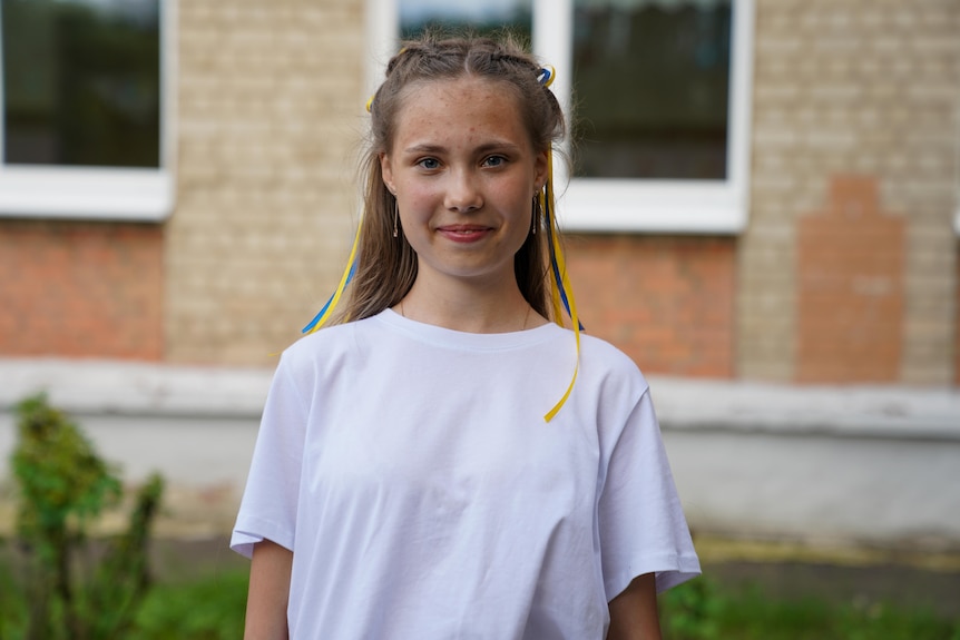 A teenage girl, her blonde hair braided away from her face with blue and yellow ribbons, smiles at the camera