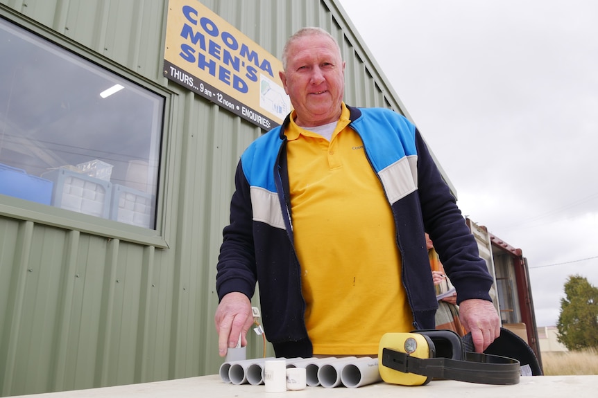 Darrell smiles at the camera, wearing a yellow polo, with PVC piping on a table and the Cooma men's shed sign in the background