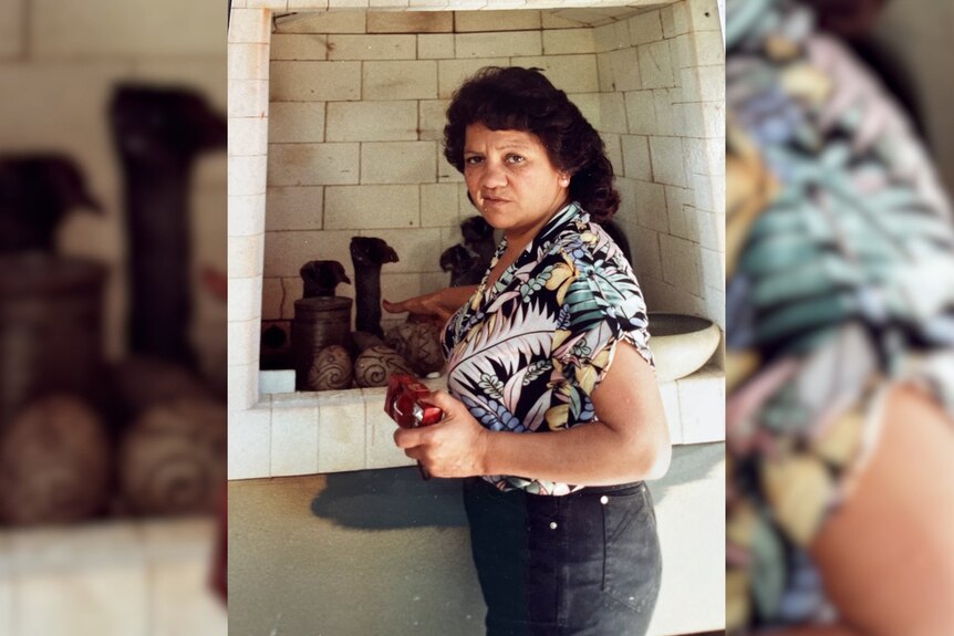 Sally looks to the camera while standing in front of a tiled kiln, her hair down in loose waves and wearing a tropical shirt.