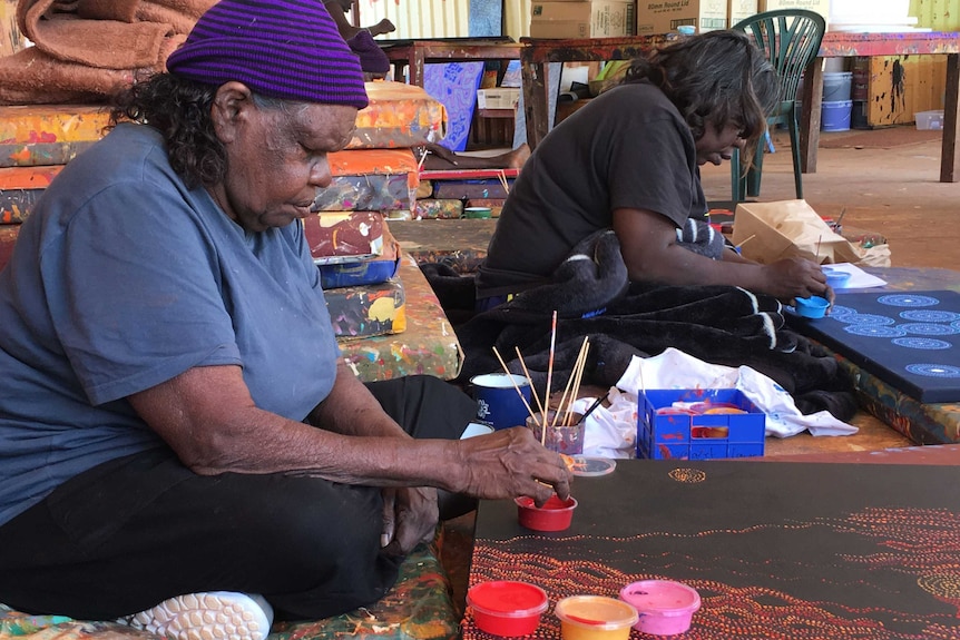 Two Indigenous artists hold paint brushes in their hands as they paint a canvas on the ground.