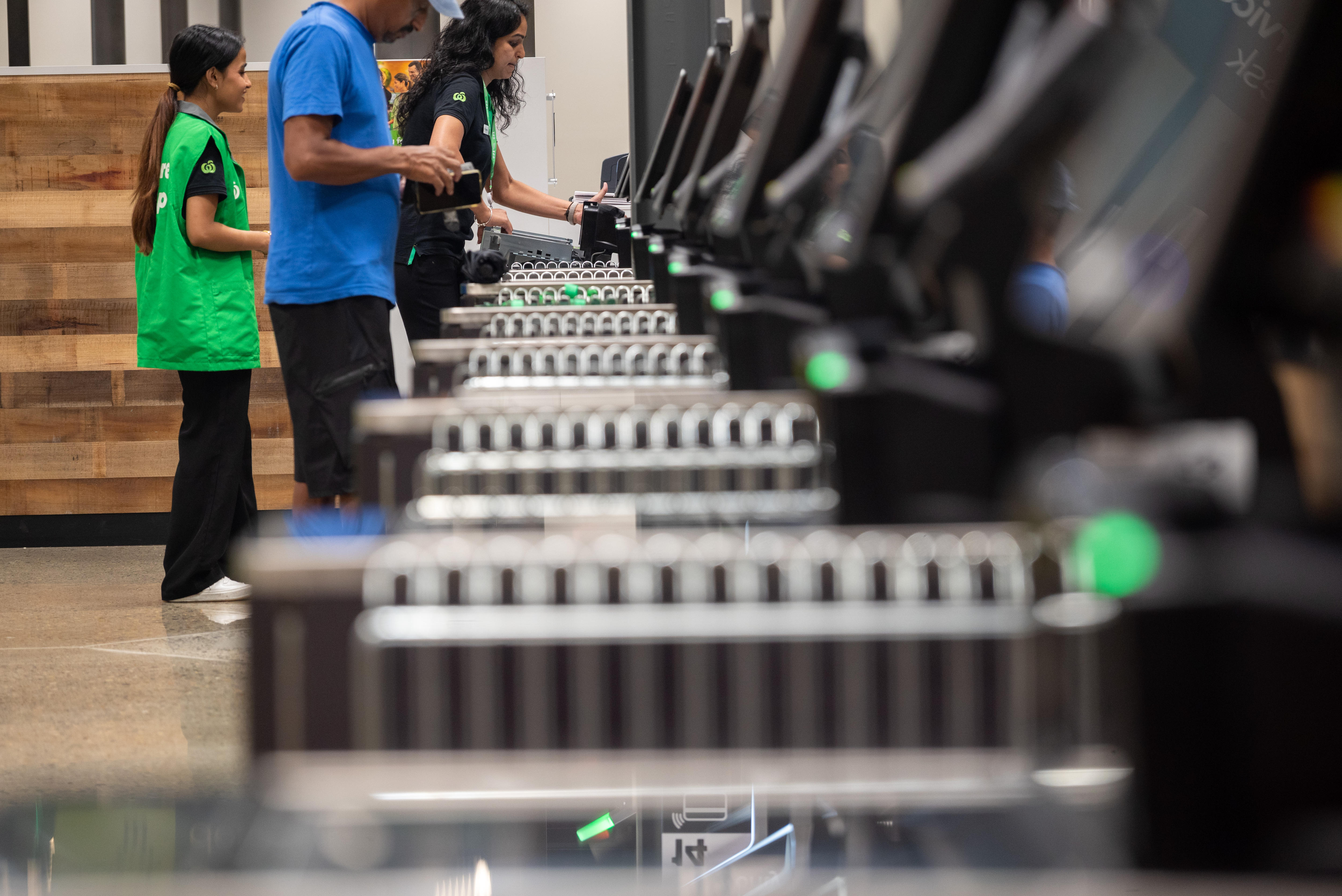 Photo taken looking down a row of self-serve checkouts to a customer and two woolworths staff.