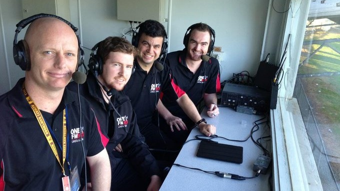 A group of men with headsets on sit at a desk overlooking a football field