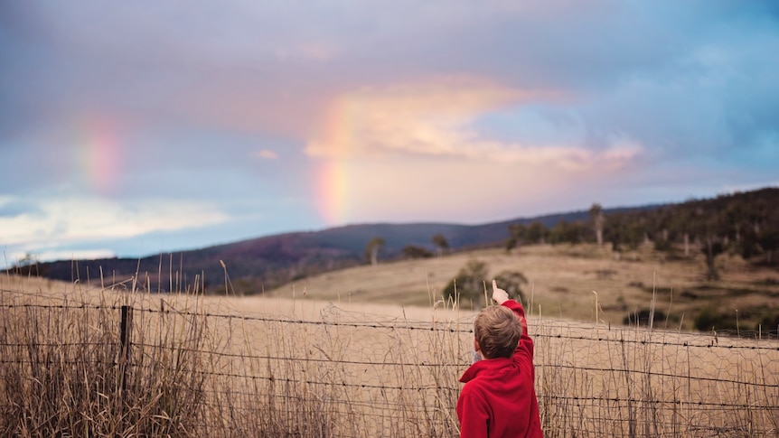 A young boy points at a rainbow beyond a fenced paddock