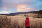 A young boy points at a rainbow beyond a fenced paddock