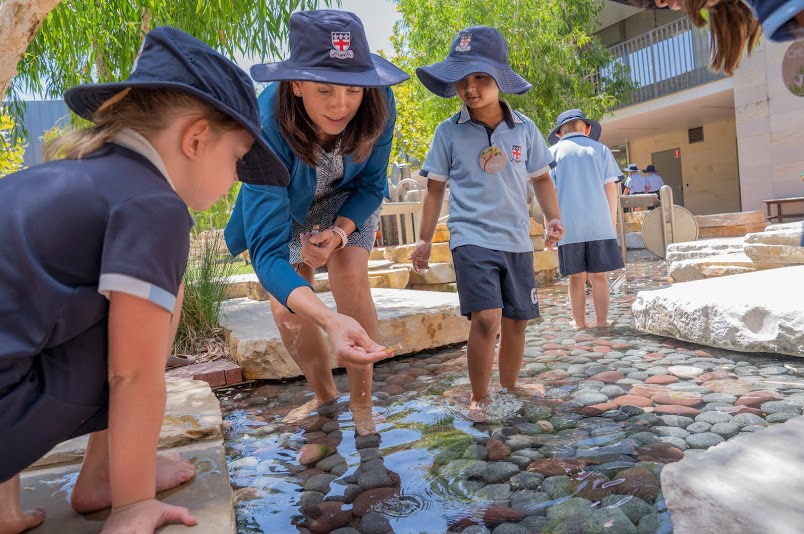 Young preparatory students and a female teacher stand in a pond looking at something closely in a school playground.