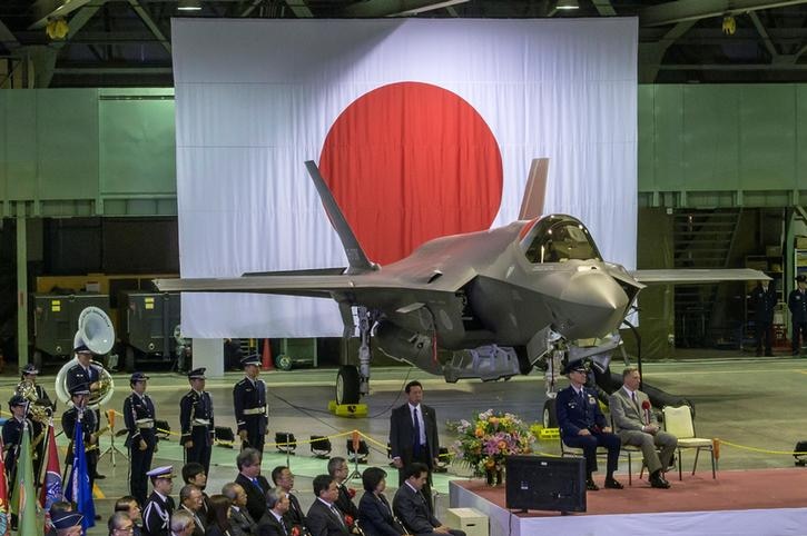 A khaki jet is seen before Japanese flag backdrop, with rows of military officials sat on chairs to the left.