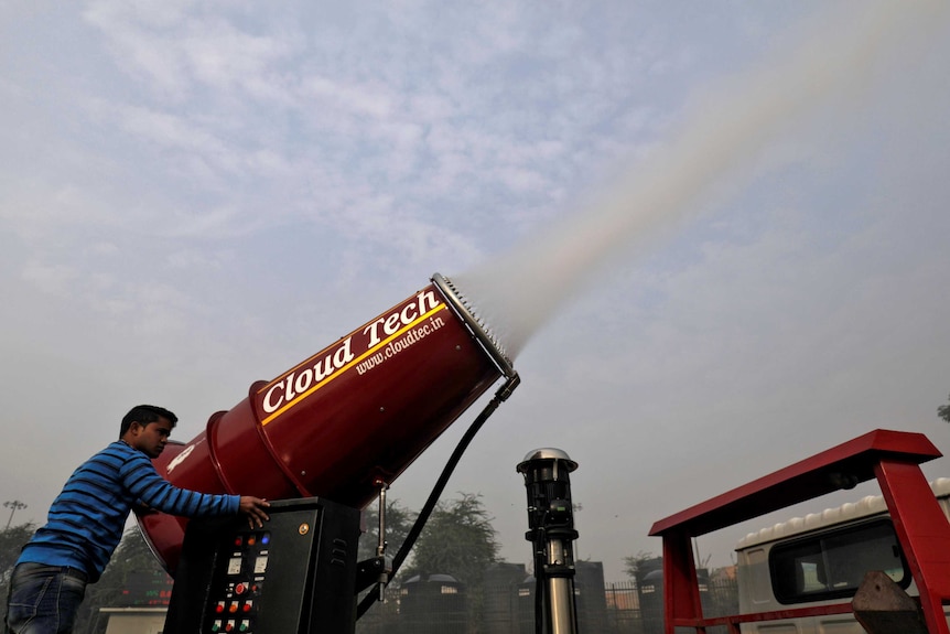 A man in a blue shirt stands outdoors behind a huge red cannon shooting what looks like dense cloud of a fine mist into the air.