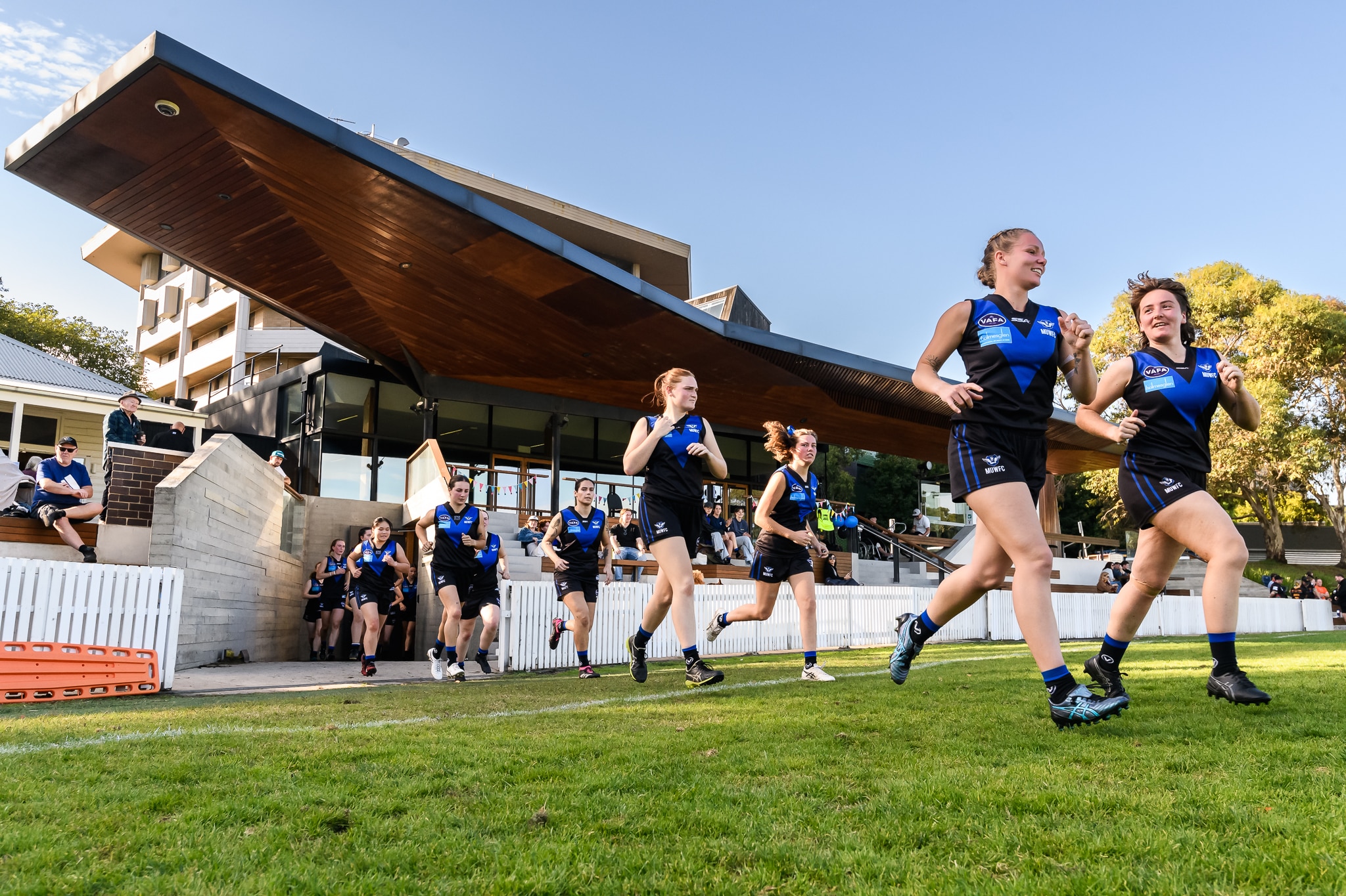Um time feminino de futebol australiano corre em direção ao campo em seu oval, com a sala do clube atrás deles. 