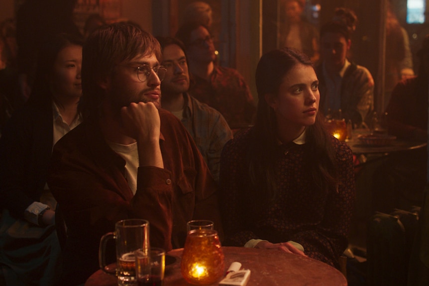 Darkened bar, with young man and woman sitting at small candle-lit table as if watching something on stage.