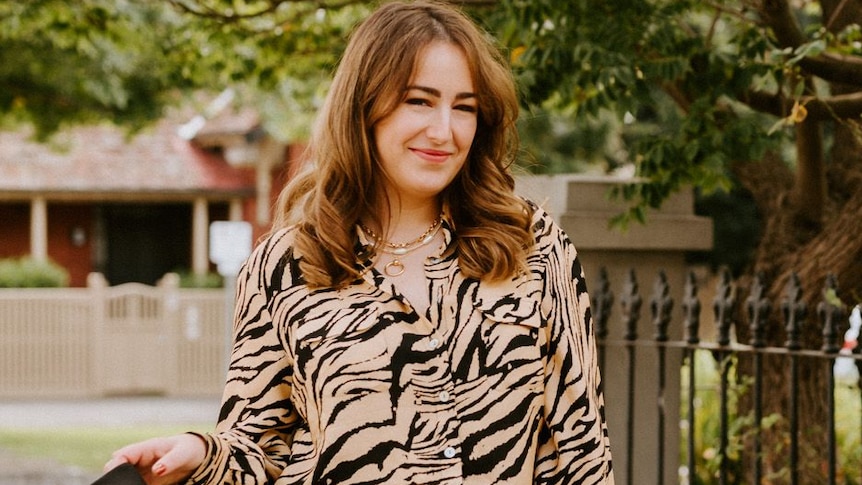 A young woman smiles and swings her jacket while standing out front of a cast-iron house fence.