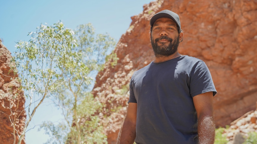 A slightly smiling Indigenous man stands in front of red rocks, looks down on camera, green trees, black t-shirt, black cap.