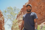 A slightly smiling Indigenous man stands in front of red rocks, looks down on camera, green trees, black t-shirt, black cap.