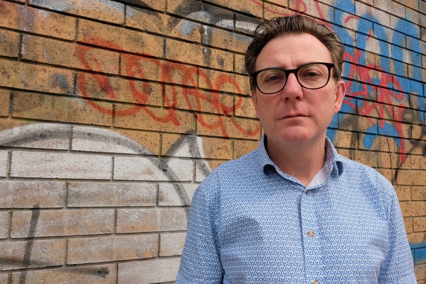 Shane Harris stands in front of a brick wall with graffiti on it.