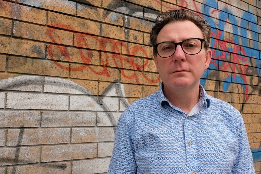 Shane Harris stands in front of a brick wall with graffiti on it.