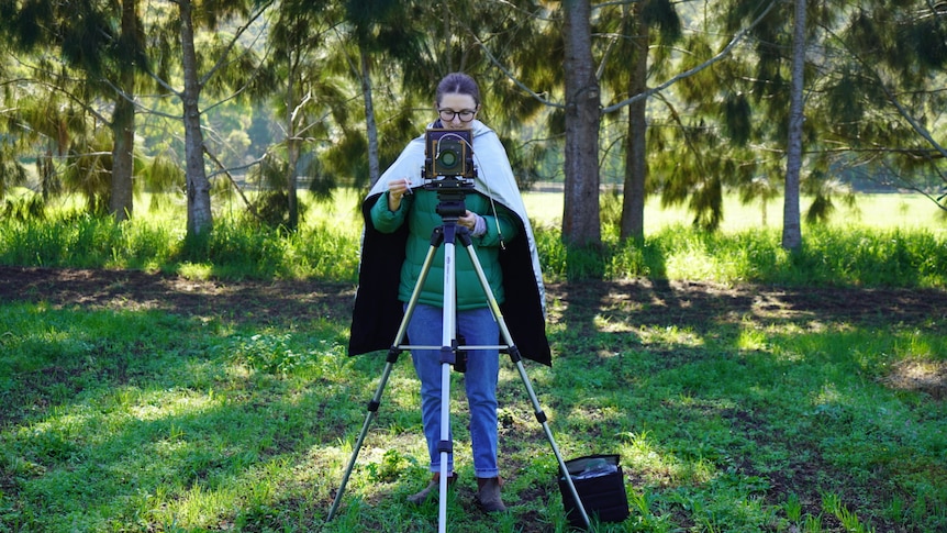 Claudia stands facing camera behind a tripod and old fashion camera in a green field