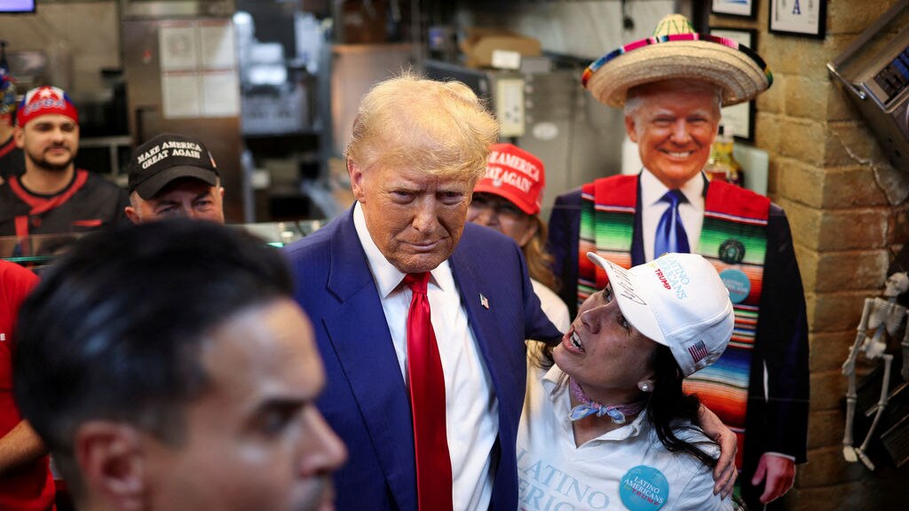Donald Trump with his arm around a woman wearing a white cap. A manequin of Donald Trump is in the background.