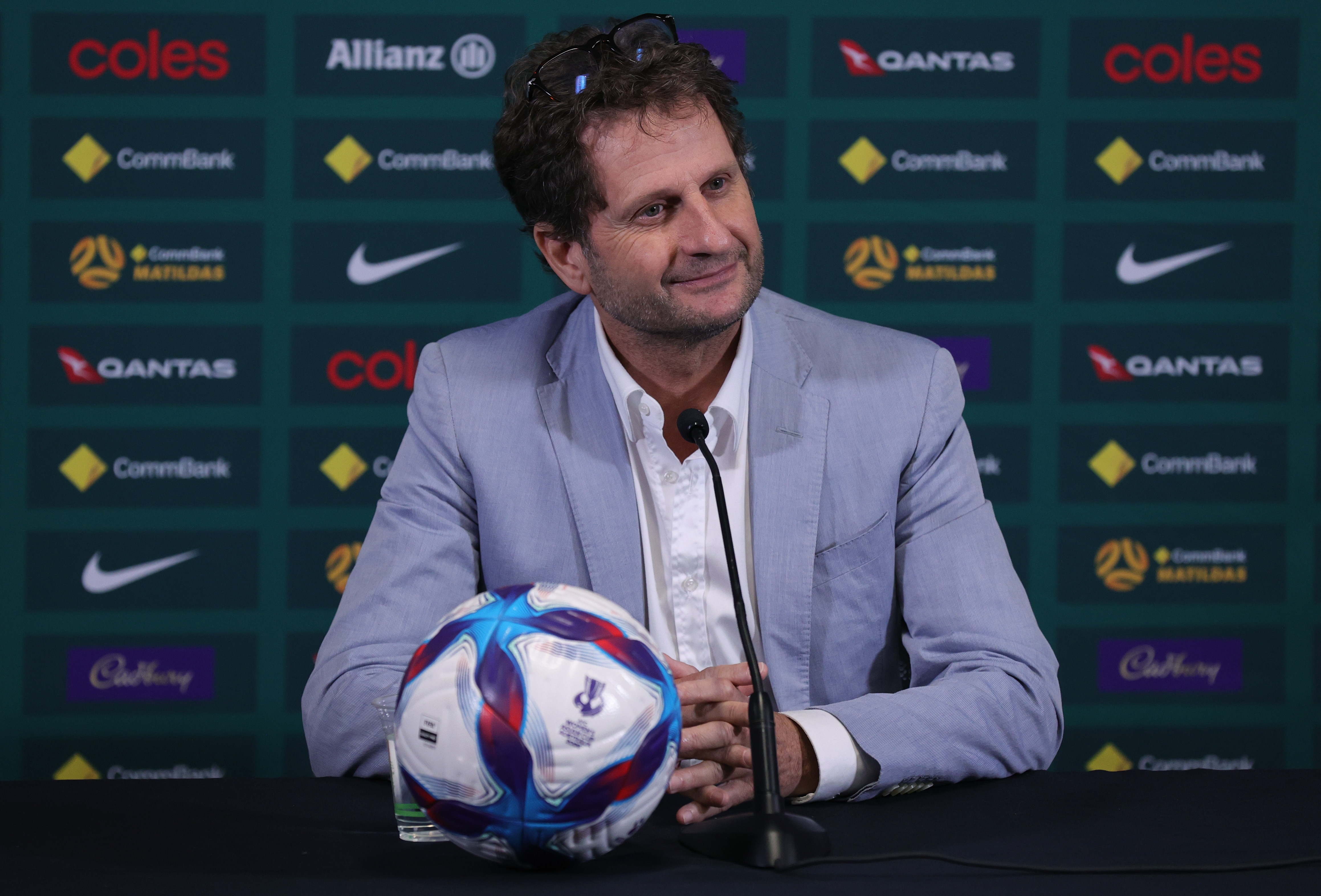 O técnico do Matildas, Joe Montemurro, sorri ao ouvir uma pergunta em entrevista coletiva. Ele está vestindo um blazer azul-cinza