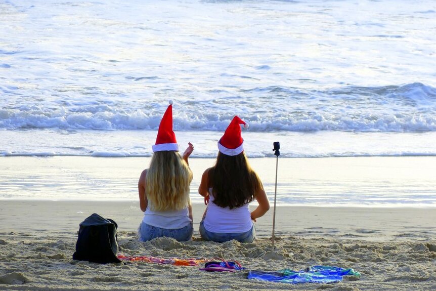 Two women sitting on the beach facing the water and wearing Santa hats