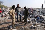 Three men, two of whom are wearing balaclavas, stand among the rubble of a destroyed village.