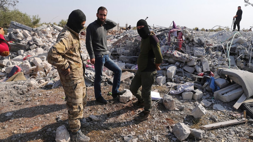Three men, two of whom are wearing balaclavas, stand among the rubble of a destroyed village.