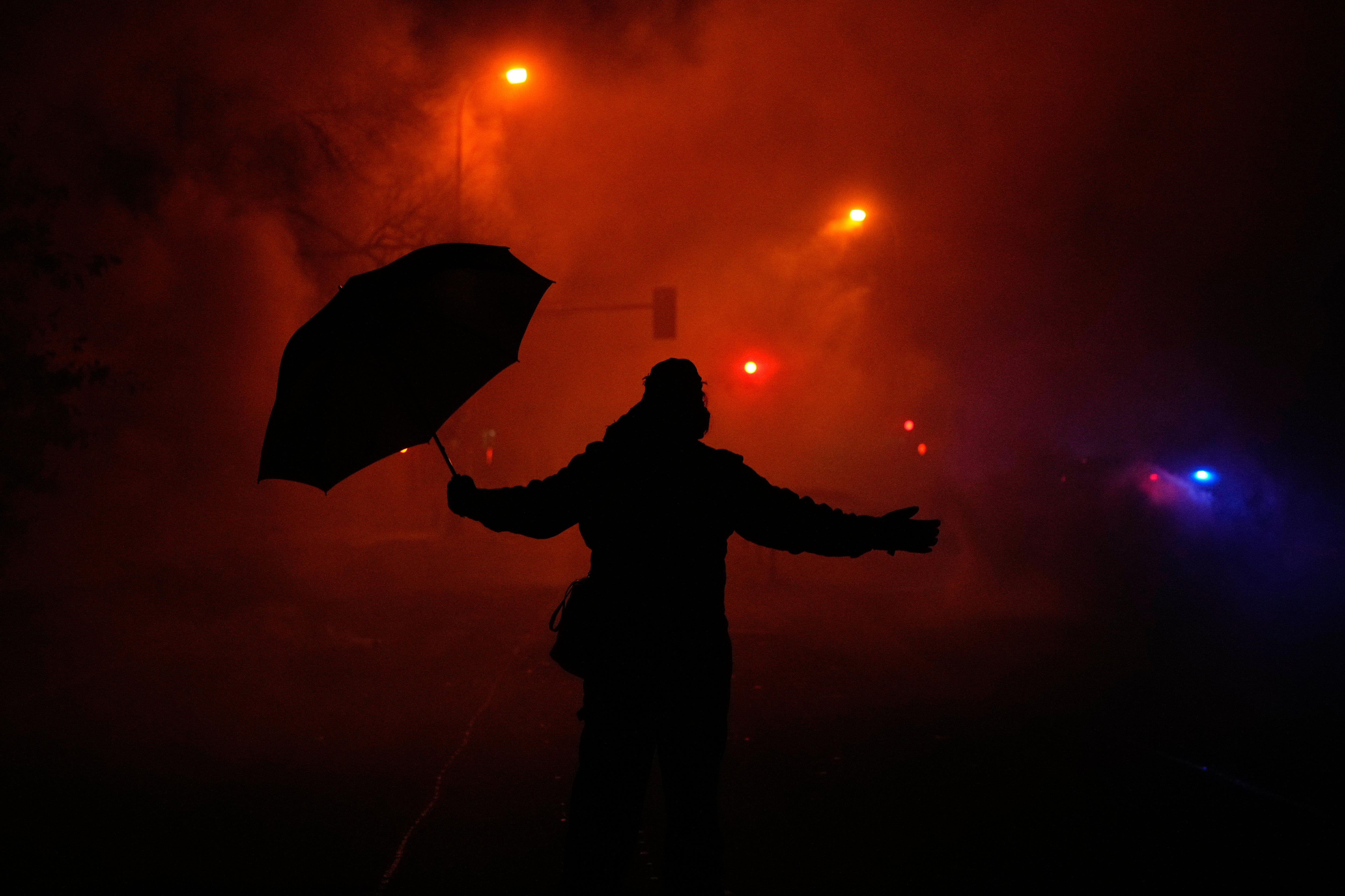 A silhueta de uma pessoa segurando um guarda-chuva sob gás lacrimogêneo em uma estrada larga à noite.