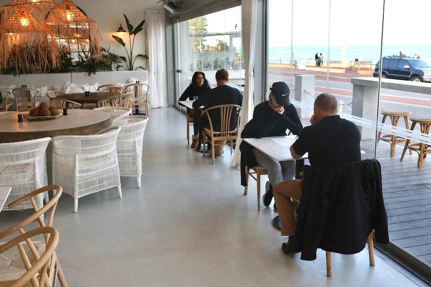Two couples sit at separate tables in a cafe next to windows along the beach.