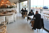 Two couples sit at separate tables in a cafe next to windows along the beach.