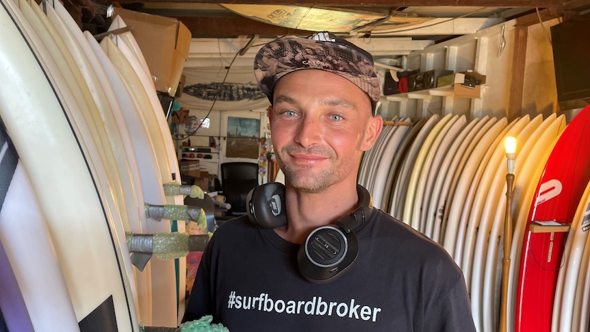 A man wearing a t-shirt and cap stands in front of a row of surfboards in a shed.