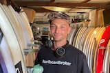A man wearing a t-shirt and cap stands in front of a row of surfboards in a shed.