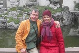 Colour photo of Kerstin Pilz sitting on a wall in the embrace of a man with short brown hair. Both wear bright puffer jackets.